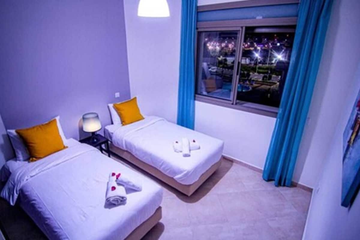 A hotel room with two beds, a nightstand, a window with curtains, and a view of the outside.