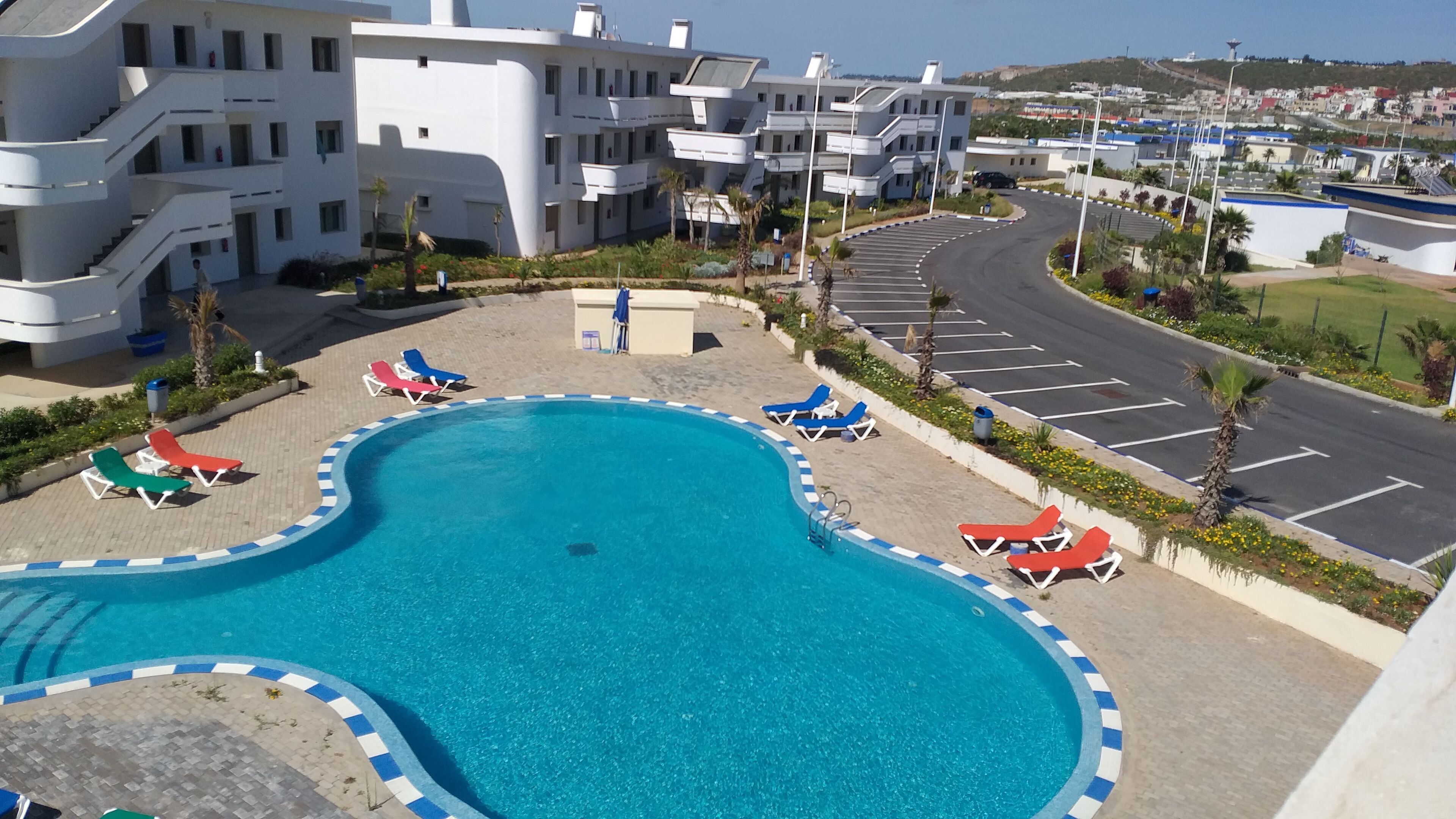Una piscina con lettini, un'area parcheggio e edifici moderni sullo sfondo.