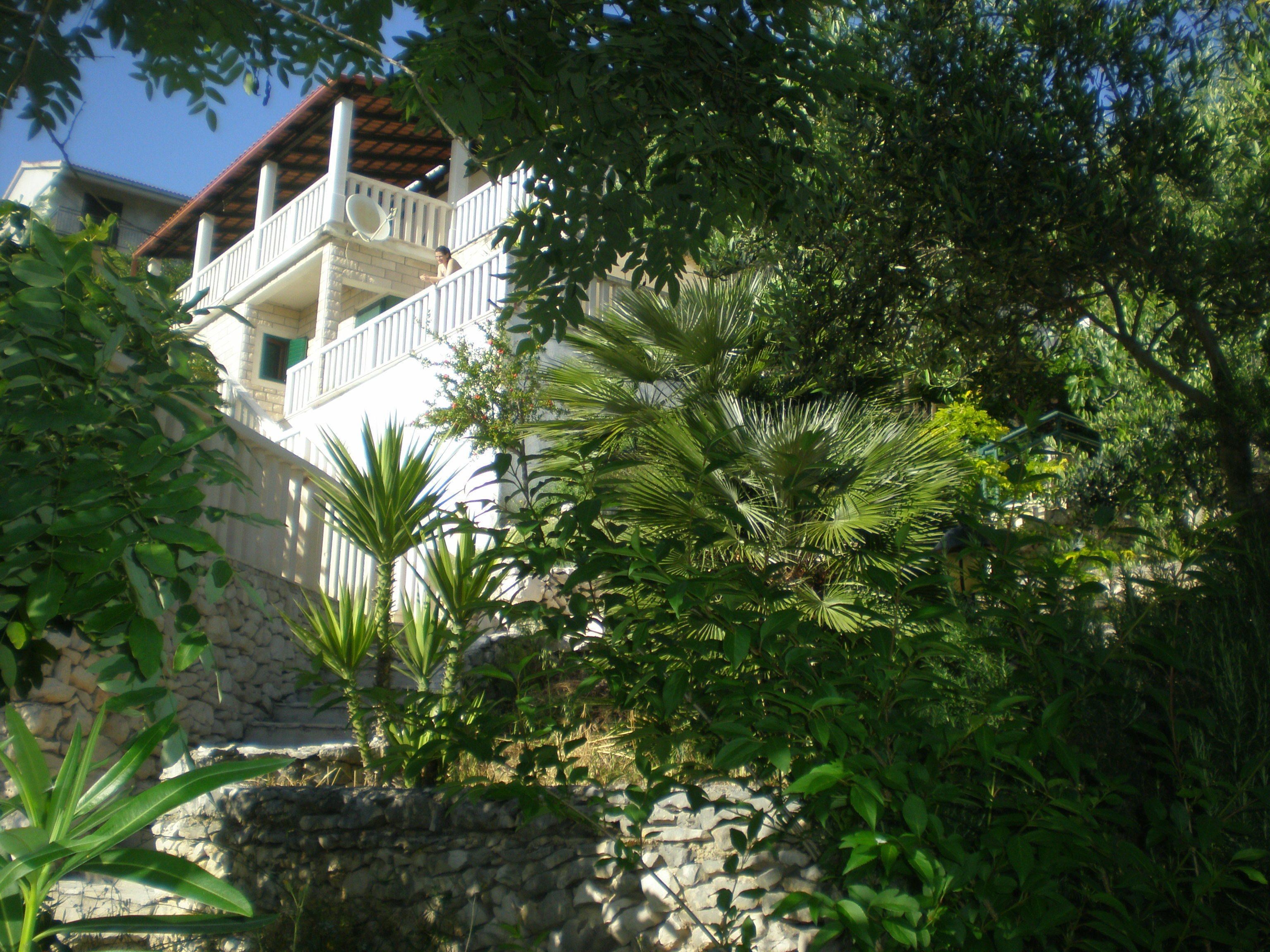 A white building with a balcony, surrounded by lush greenery and a stone wall.