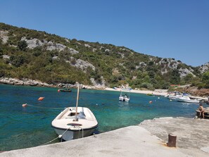 A coastal scene with boats docked at a pier, clear turquoise water, and a rocky hillside in the background.