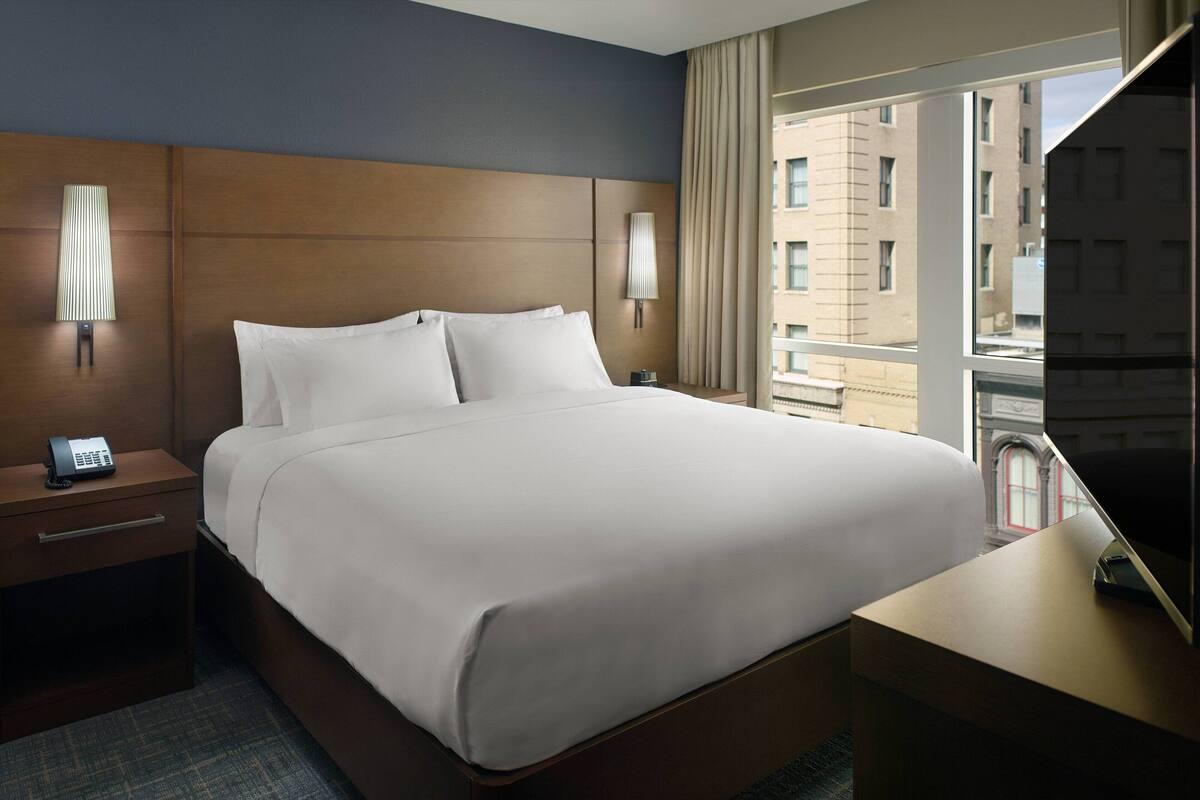 Een hotelkamer met een groot bed, bedlampen, een bureau met een televisie en een raam met uitzicht op de stad.