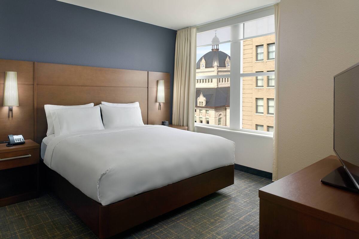 Hotelkamer met een groot bed, een nachtkastje met telefoon, een bureau met laptop en een raam met uitzicht op een gebouw.