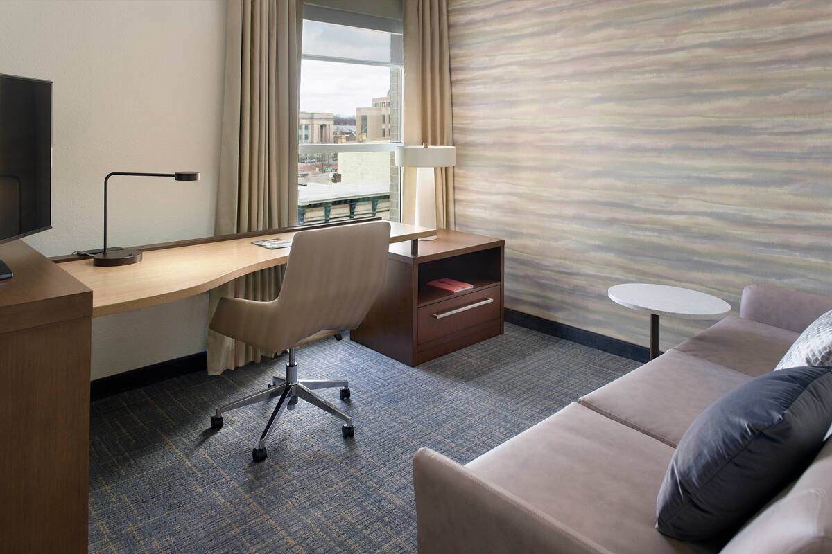 Een hotelkamer met een bureau, stoel en een bank. Op het bureau staat een televisie, een lamp en een raam met gordijnen.