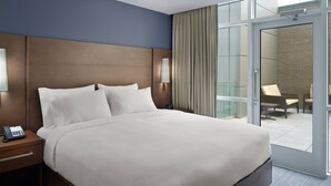 Quarto de hotel com uma cama grande, telefone de apoio, luminária e vista para uma área de estar externa através de uma porta de vidro.