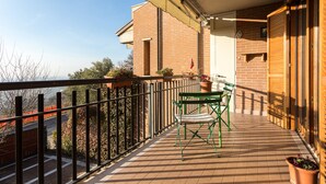 A balcony with a green chair and table, potted plants, and a view of the street and trees. - Holiday rental apartment Le Fate di Campagna (Assisi)