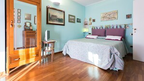 A bedroom with a wooden dresser, a bed with floral bedding, and a wall-mounted shelf with various items. - Holiday rental apartment Le Fate di Campagna (Assisi)