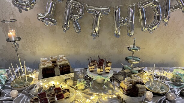A birthday dessert table with various cakes and sweets, illuminated by string lights. - Hotel Carina (Tczew)