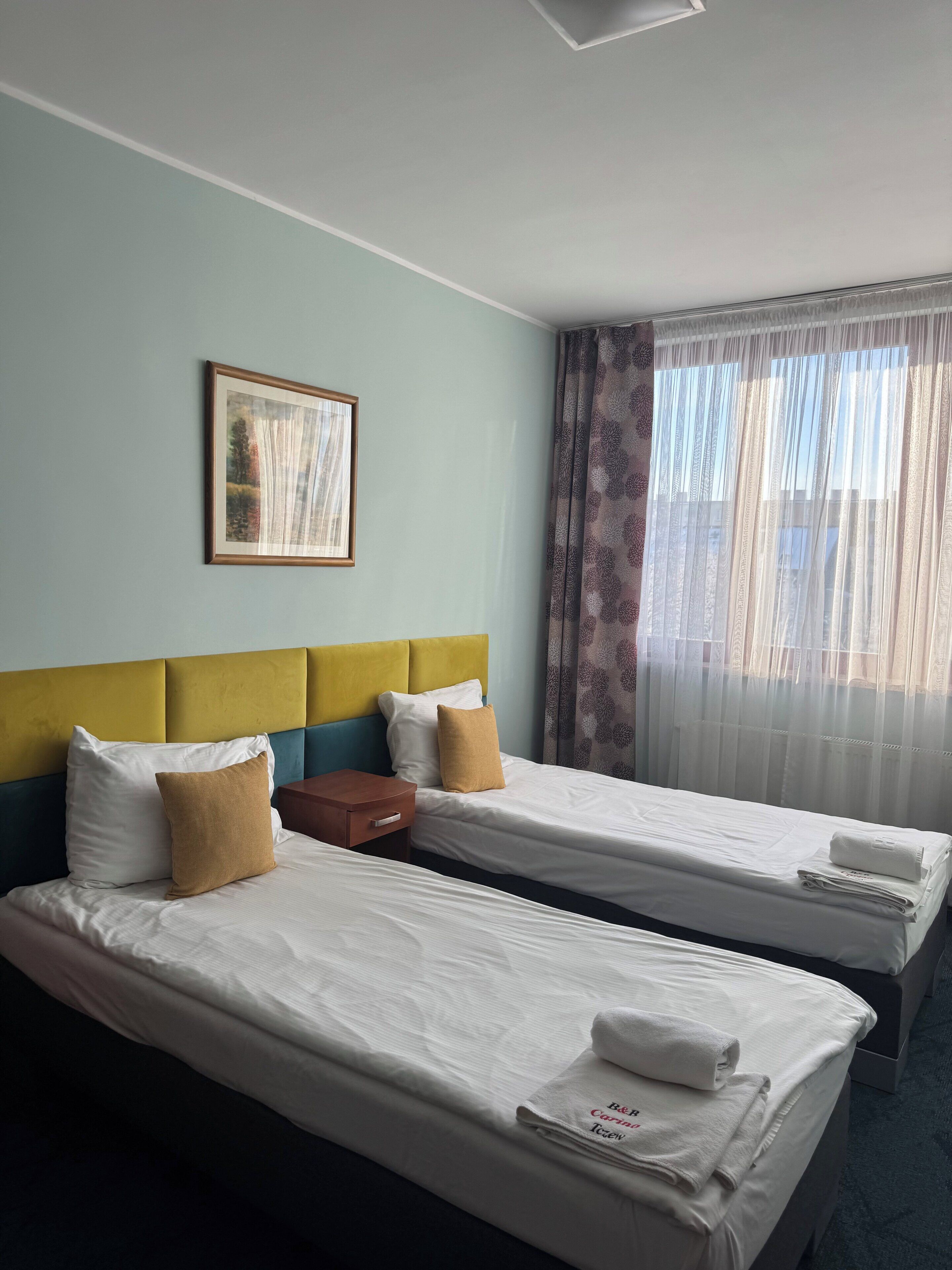 Een hotelkamer met twee bedden, een lijst met een foto aan de muur en dunne gordijnen voor het raam.