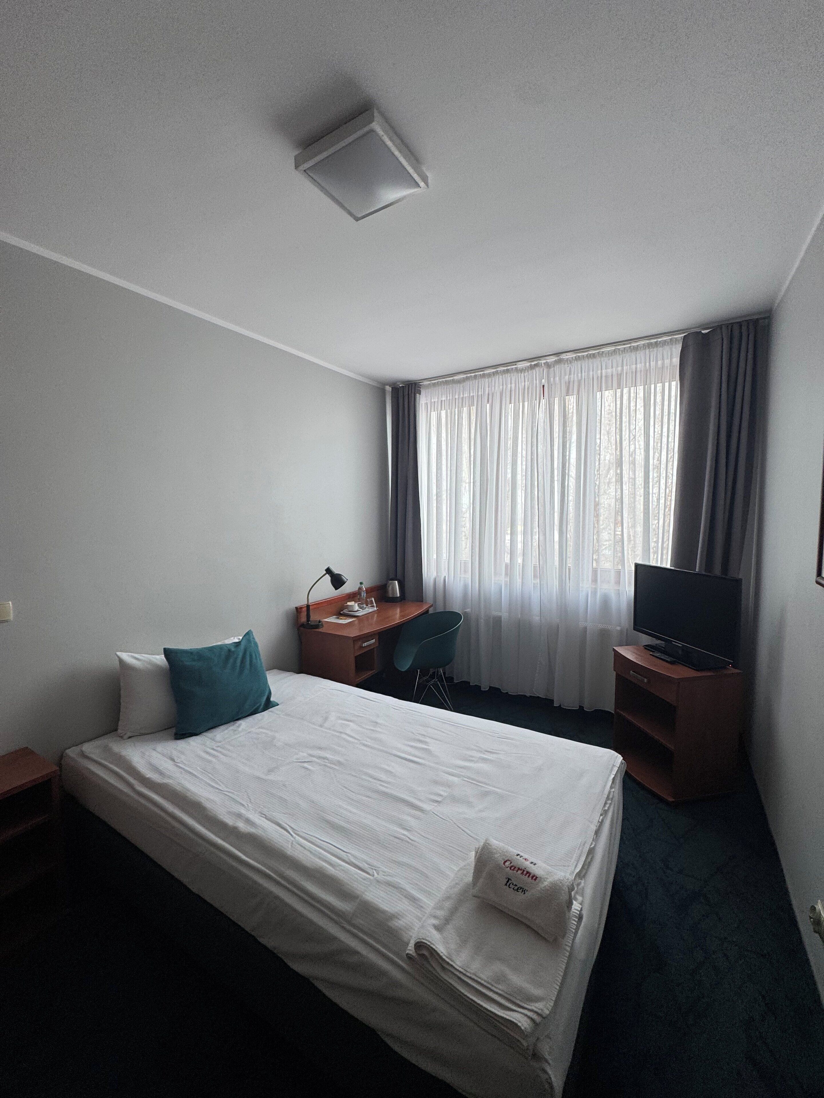 Una habitación de hotel con cama, escritorio, silla, televisor y ventana con cortinas.