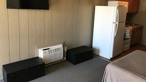 Habitación compacta con televisor de pantalla plana en la pared, refrigerador blanco, kitchenette pequeña con armarios de madera y una maleta negra en el suelo.