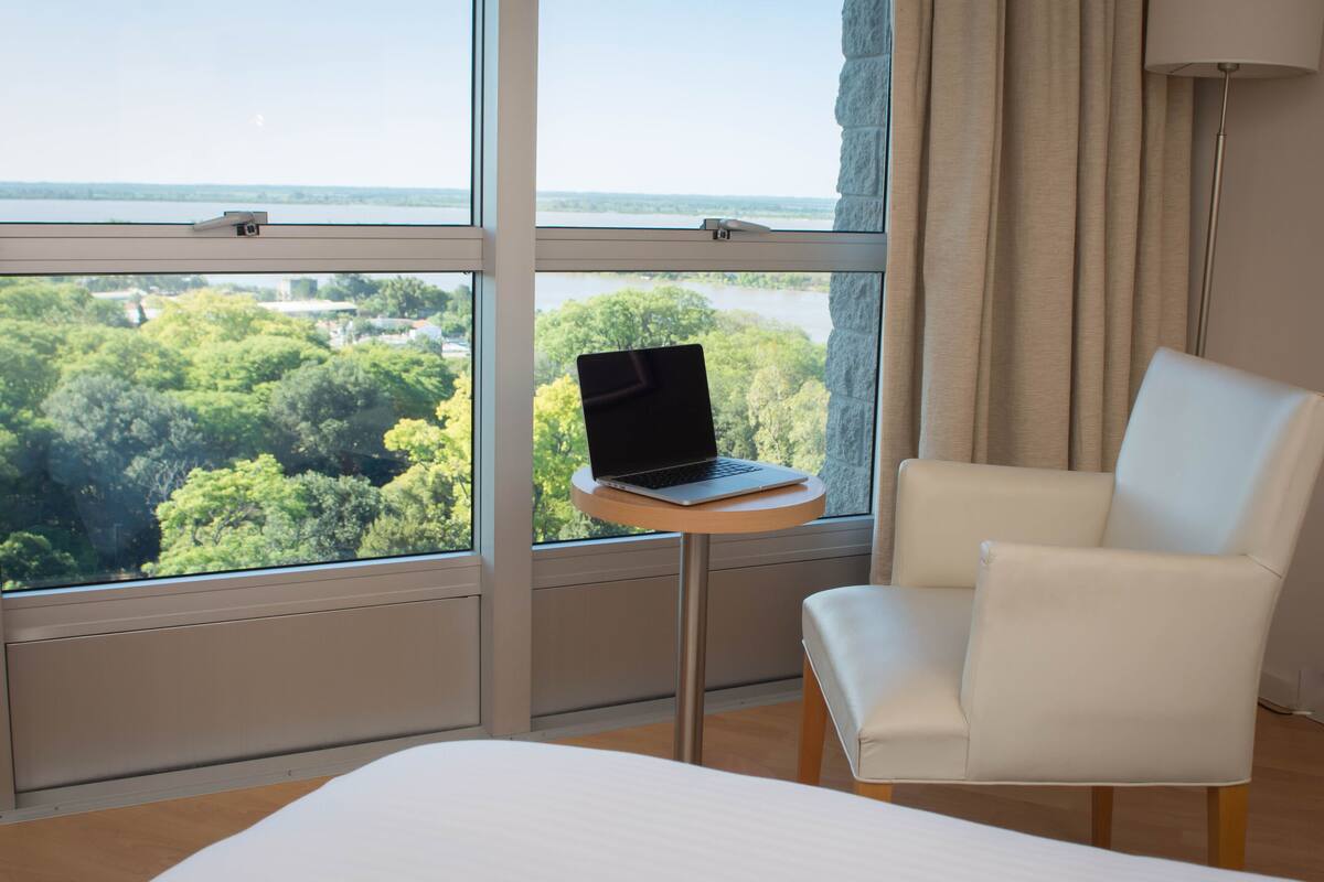 Une chambre d’hôtel avec un lit, une chaise blanche, une petite table ronde avec un ordinateur portable, et une grande fenêtre donnant sur un lac et des arbres.