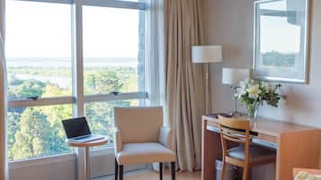 Een hotelkamer met een groot raam, een bureau met een laptop, een stoel, een lamp en een vaas met bloemen.