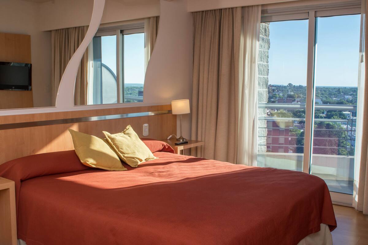 Une chambre d’hôtel avec un grand lit, un téléviseur fixé au mur, une table de chevet et une vue sur la ville grâce à de grandes fenêtres.
