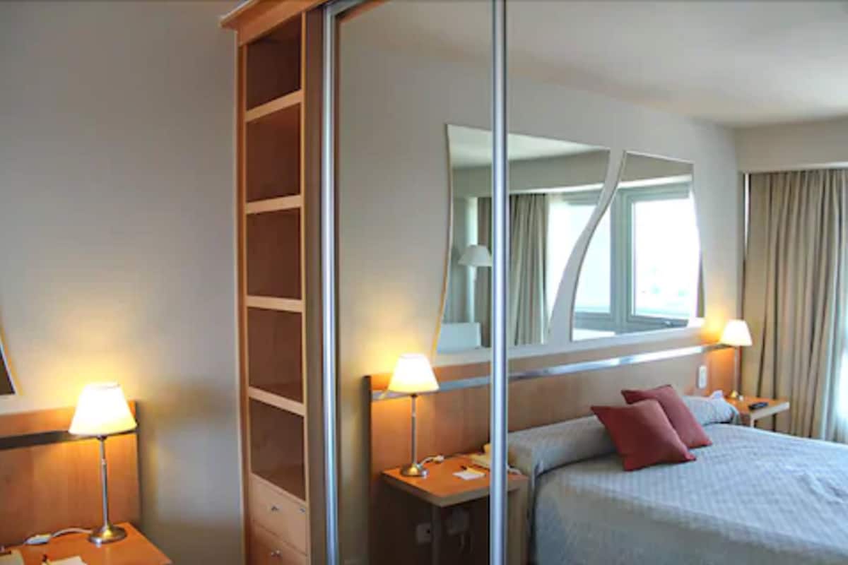 Une chambre d’hôtel avec un grand miroir, deux tables de chevet avec des lampes, un lit avec un oreiller rouge et une armoire.