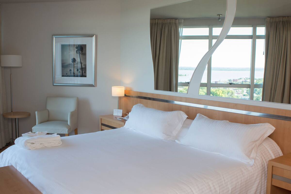 Une chambre d’hôtel avec un grand lit, une chaise, une table de chevet, une lampe et un tableau encadré au mur.