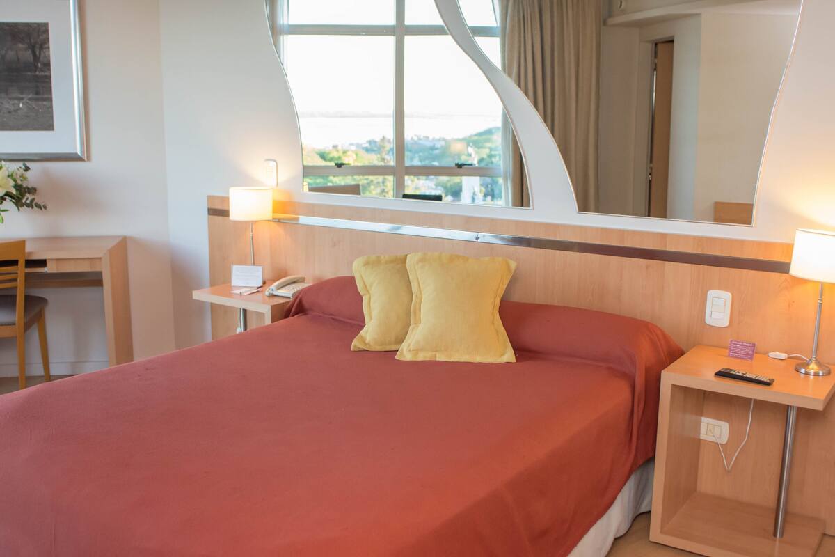 Une chambre d’hôtel avec un lit, une table de chevet, une lampe et une fenêtre donnant sur un paysage.