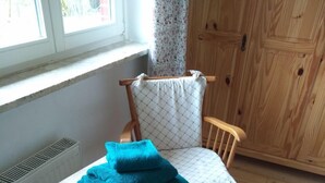 A room with a wooden chair, a small table with a folded blue towel, a patterned rug, and a window with a view of greenery.