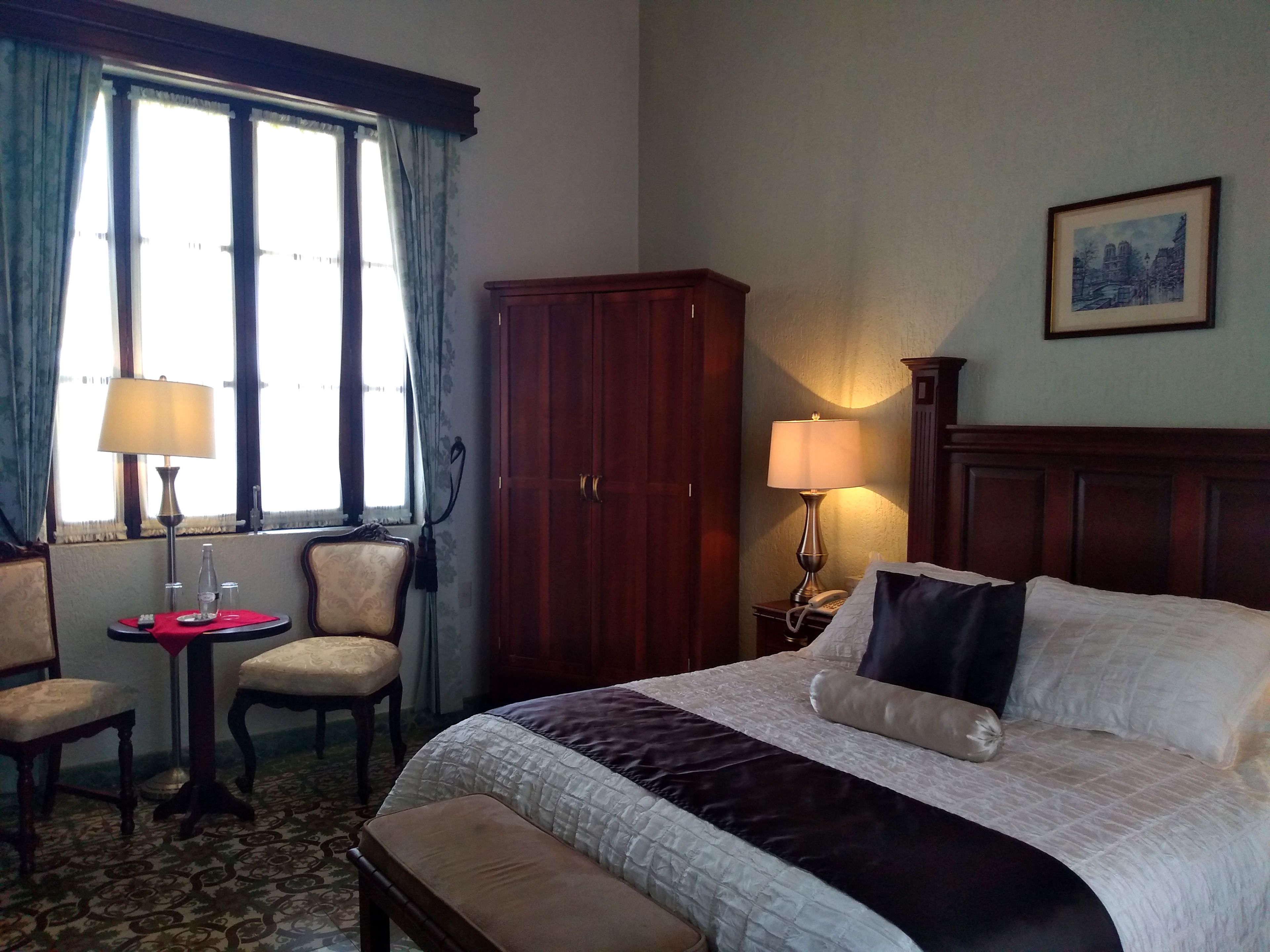 Une chambre d’hôtel comprenant un lit, deux chaises, un bureau, une lampe, une armoire et un tableau encadré au mur.