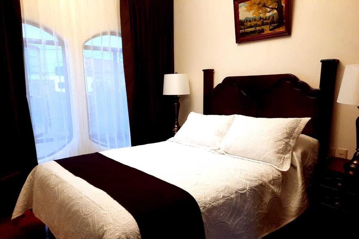 Een netjes opgemaakt bed met een witte dekbedovertrek en een donkere plaids, een bedlampje en een schilderij aan de muur.