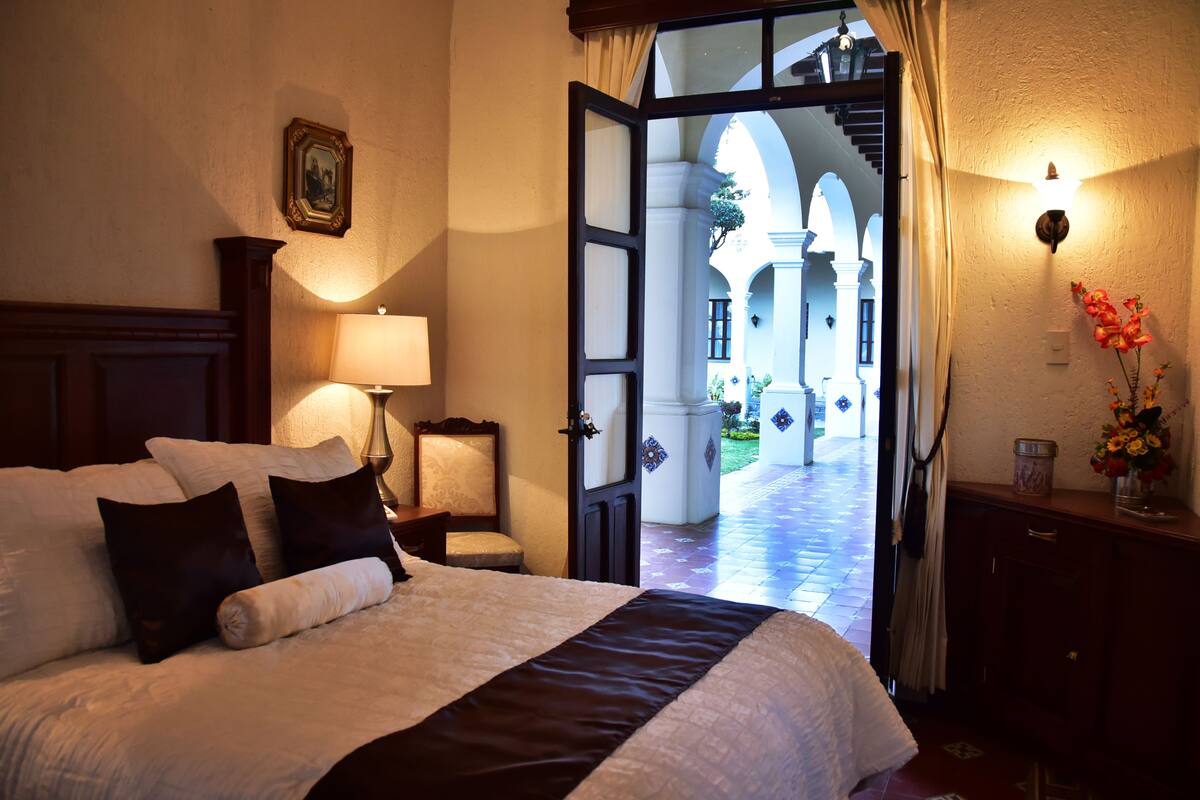 Een hotelkamer met een bed, nachtkastje, lamp, stoel en uitzicht op een open binnenplaats door een geopende deur.