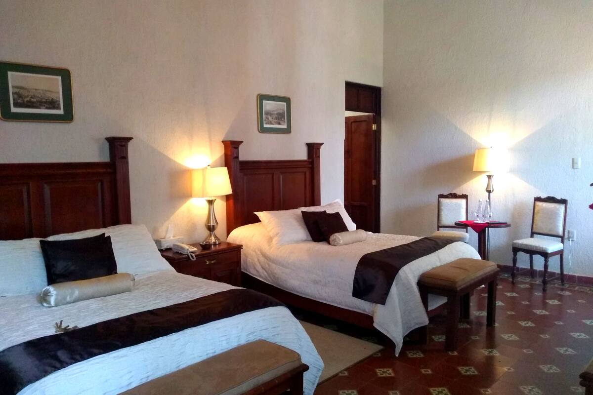 Een hotelkamer met twee bedden, een nachtkastje met een lamp, een stoel en een kleine tafel met glazen.