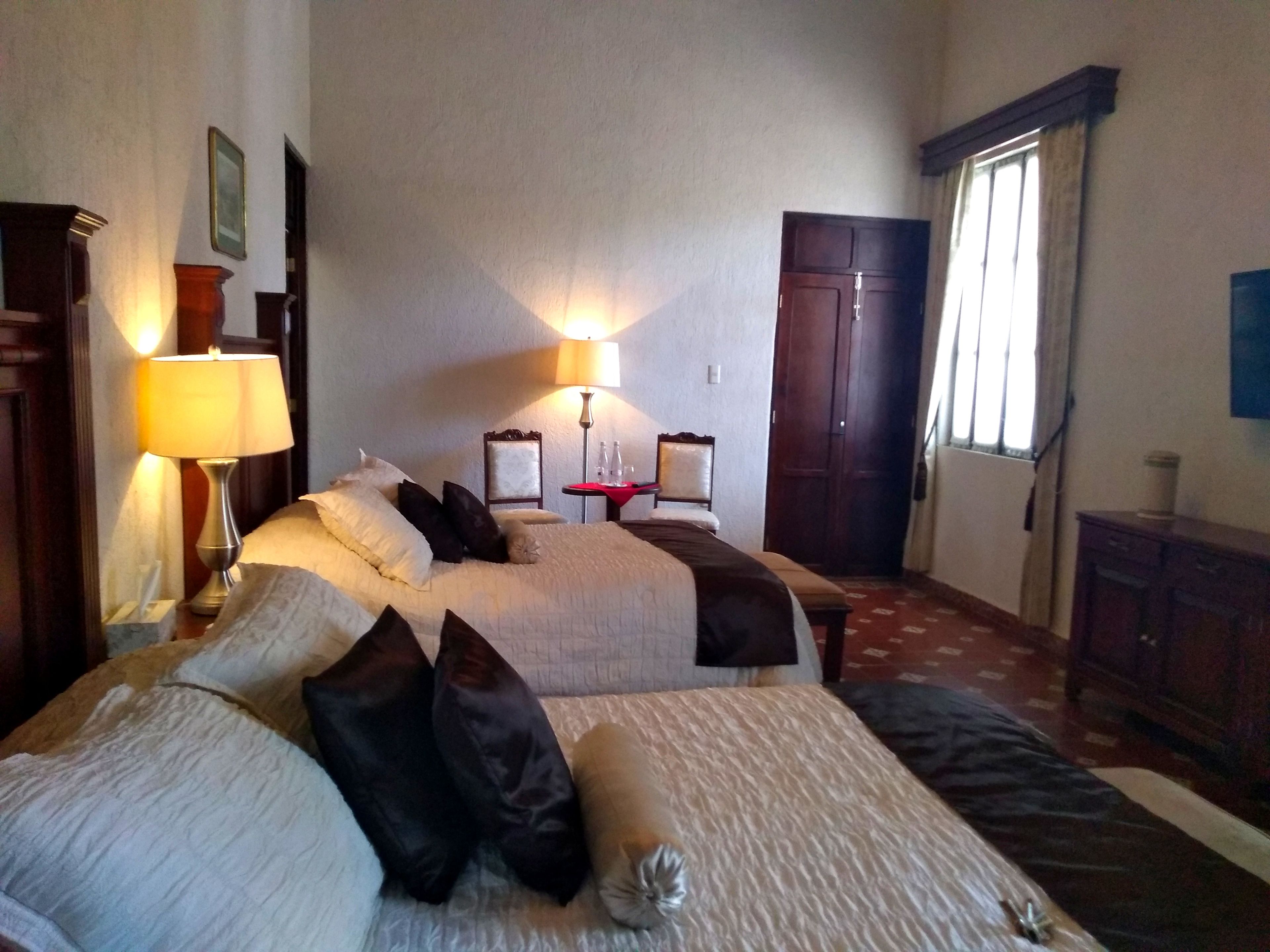 A hotel room with two beds, a nightstand with a lamp, a small table with a red tablecloth, a chair, and a wardrobe.