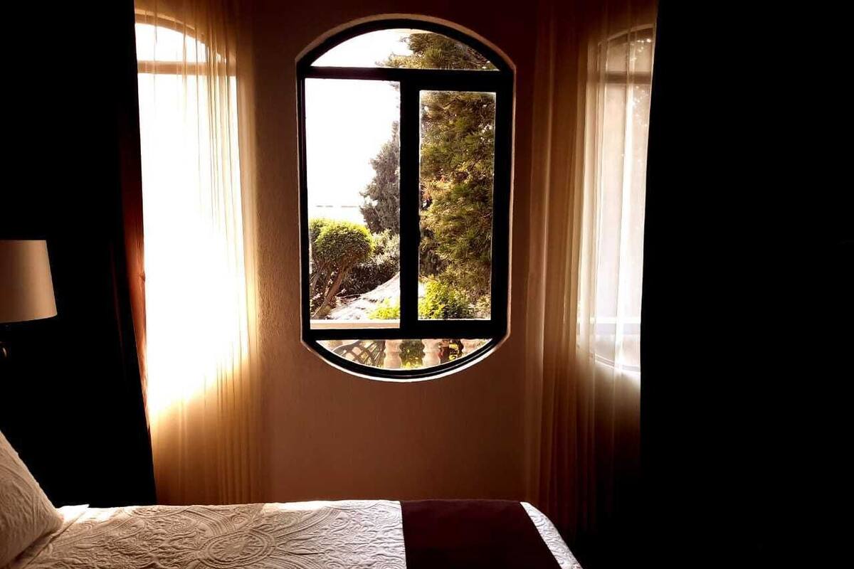 Een slaapkamer met een bed, een raam met uitzicht op groen, en gordijnen.
