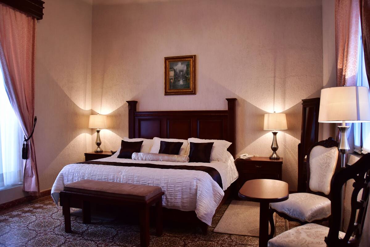 Een hotelkamer met een groot bed, twee nachtkastjes met lampen, een stoel, een kleine tafel en een schilderij aan de muur.