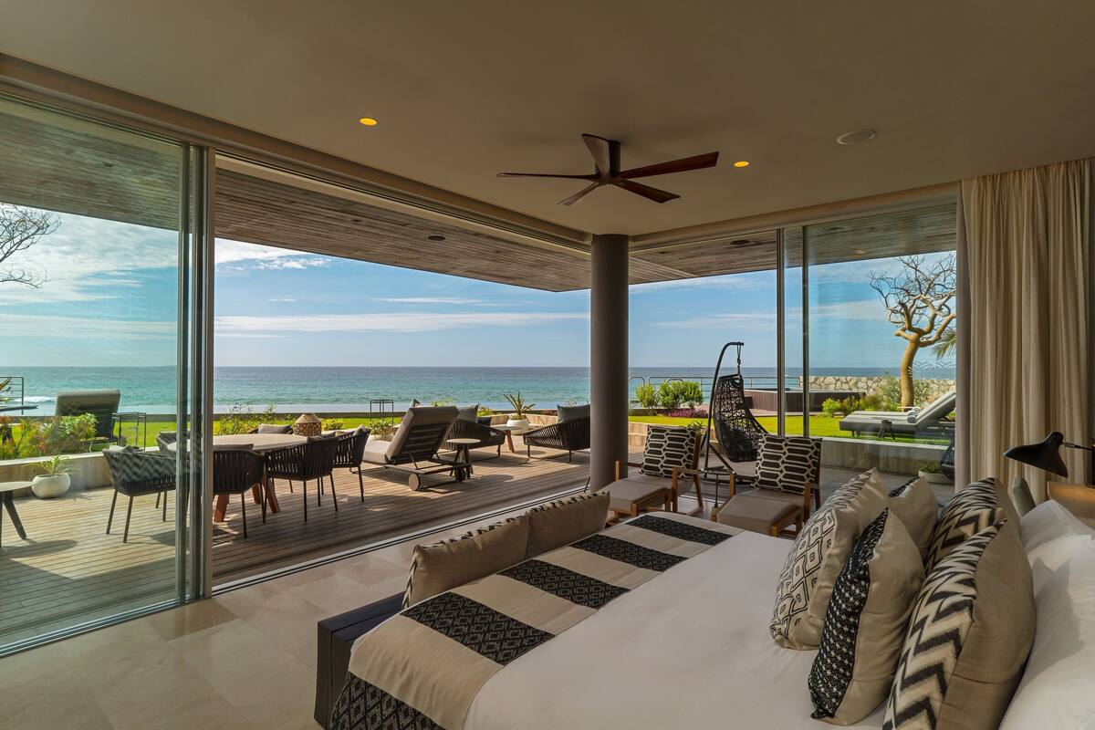 A modern bedroom with a large bed, a ceiling fan, and a view of the ocean.