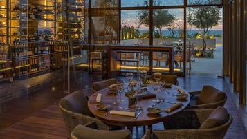 Un restaurante con mesa de madera dispuesta para comer, rodeado de sillas de mimbre y grandes ventanales con vistas al exterior.