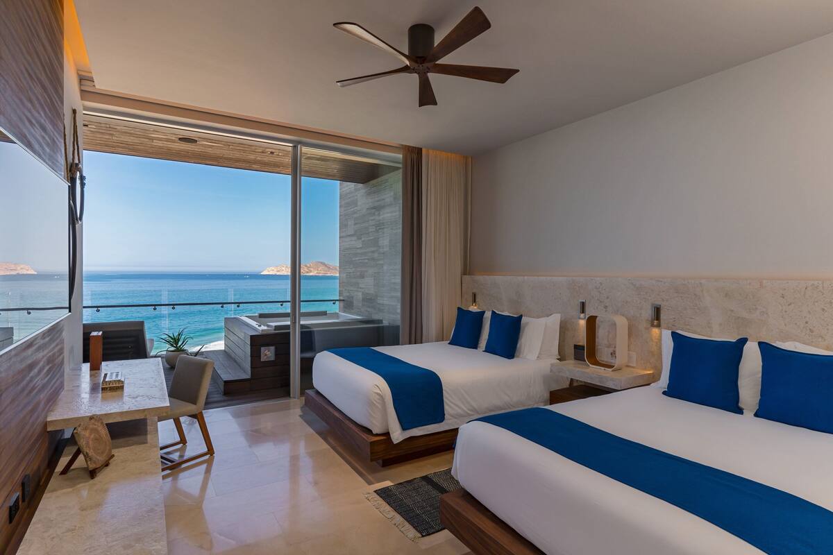 A modern hotel room with two beds, a balcony with a view of the sea, and a ceiling fan.