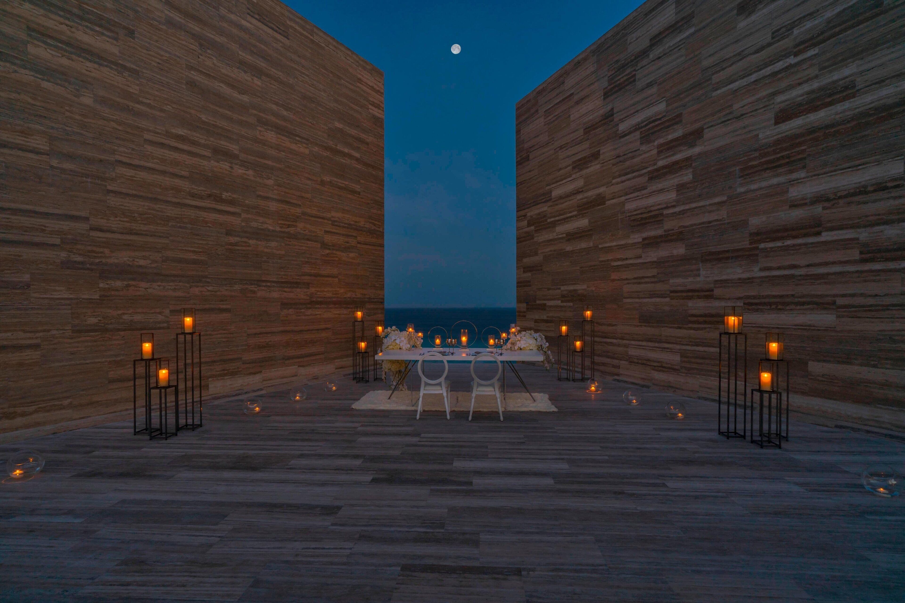 An outdoor dining area with a table set for two, flanked by tall wooden walls, under a night sky with a visible moon.