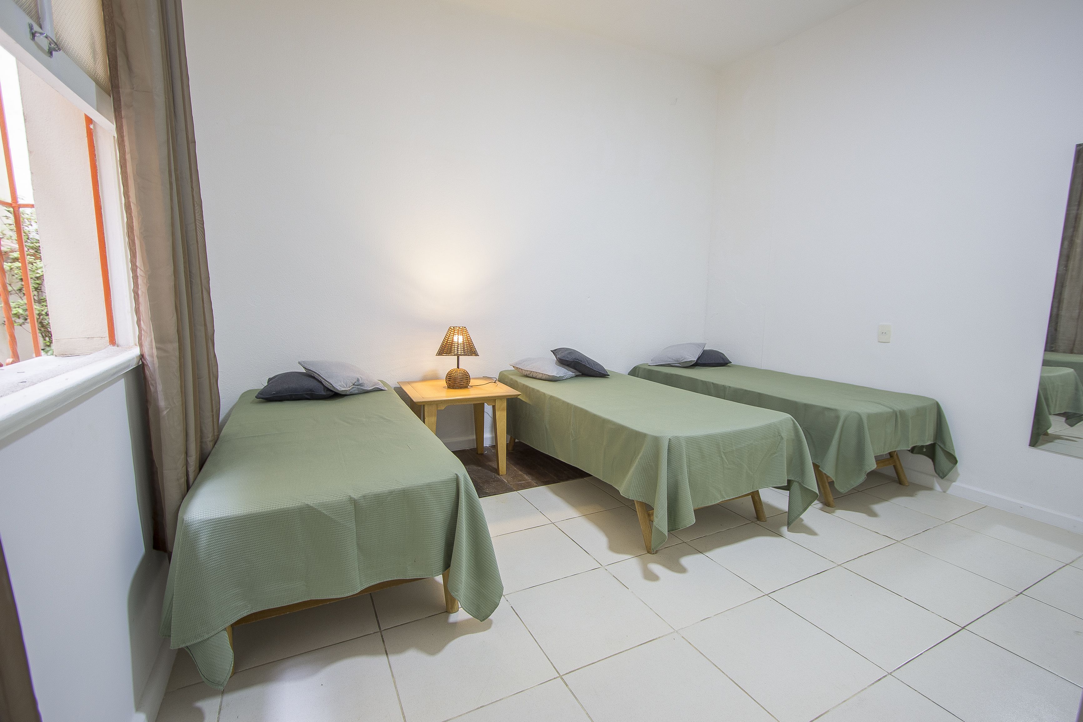 A room with three single beds, a small table with a lamp, and a window with curtains.