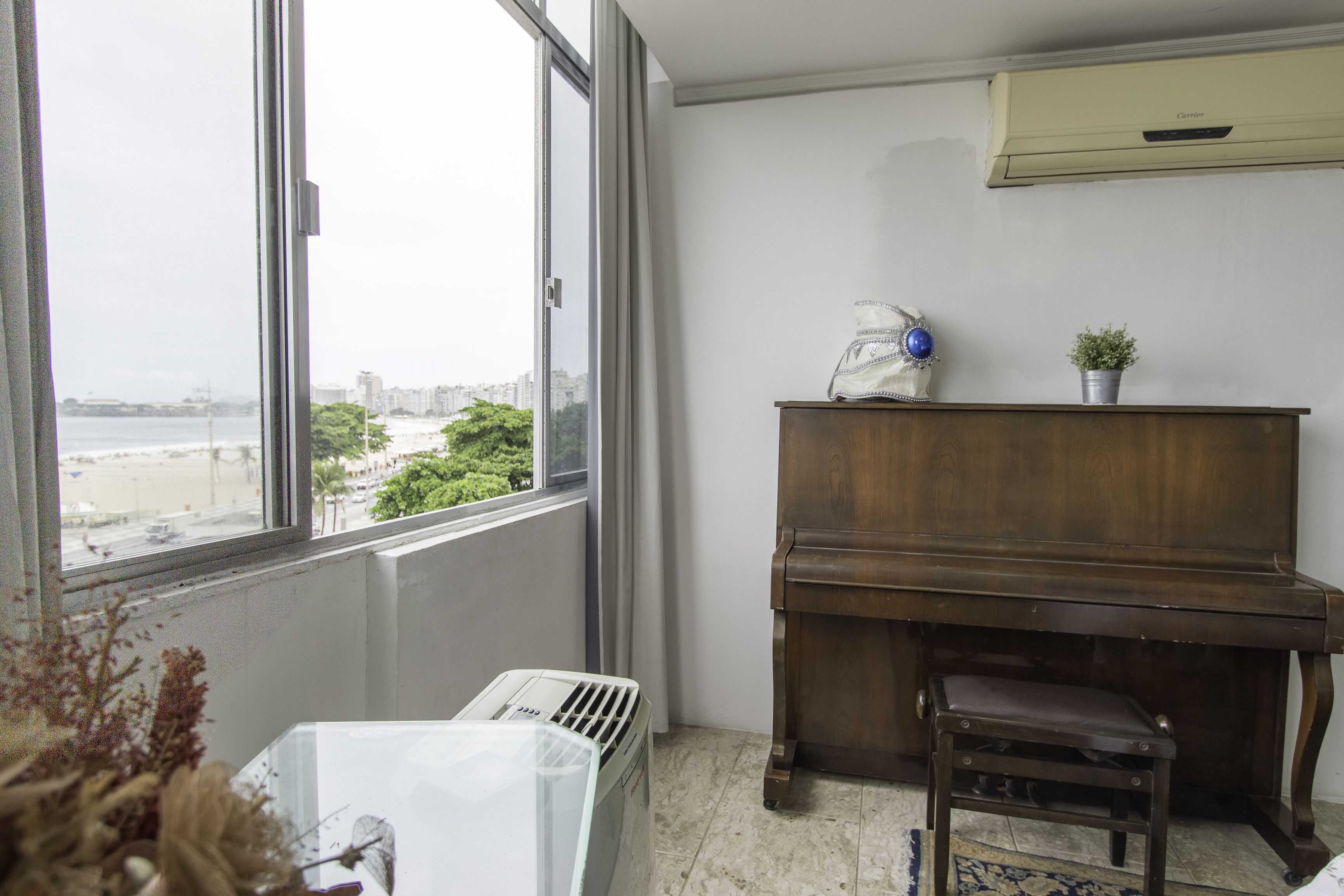 Een kamer met een piano, een radiator en uitzicht op een stadsbeeld door grote ramen.