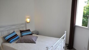 A single bed with a white metal frame, two pillows, a bedside table, and a wall-mounted light.