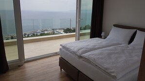 Een slaapkamer met een groot bed, uitzicht op zee door een schuifdeur en een balkon met zicht op boten en steigers.