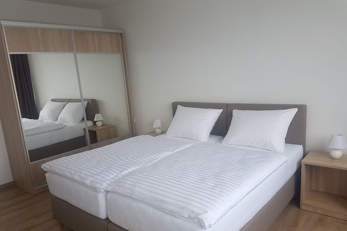Una cama bien tendida con sábanas blancas, un espejo grande y mesitas de noche con lámparas.