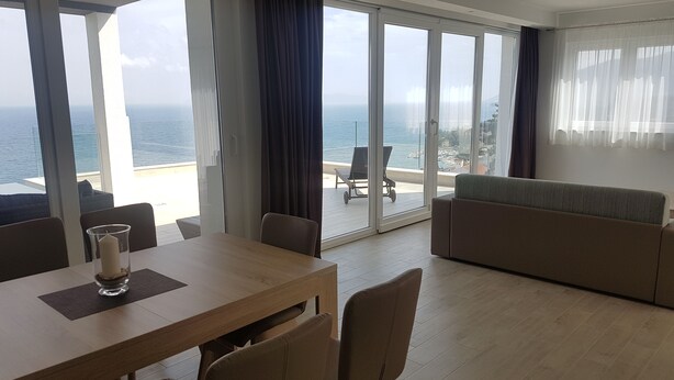 Een moderne woonkamer met een houten eettafel, een bank en uitzicht op zee door grote ramen.