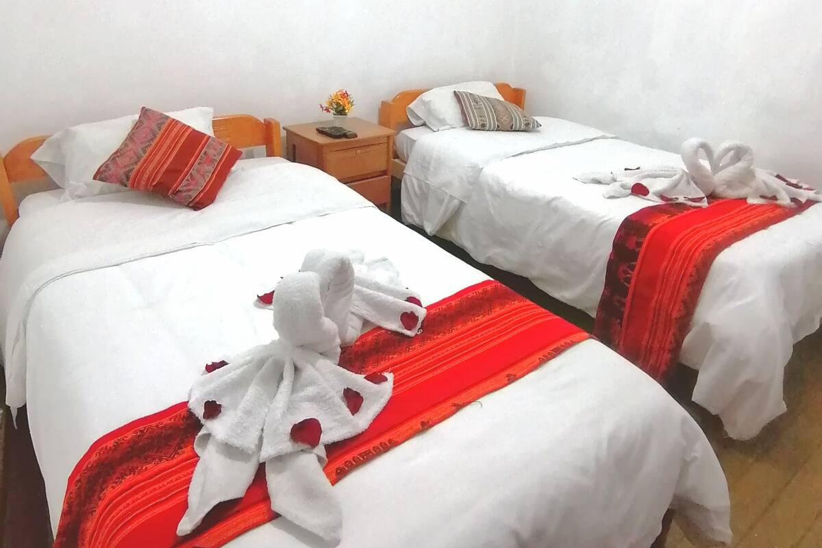 Ein Hotelzimmer mit zwei Betten, beide mit wei ßer Bettwäsche und roten Akzenten. Es gibt Nachttische aus Holz mit Lampen und einen kleinen Tisch mit einer Blumenvase.
