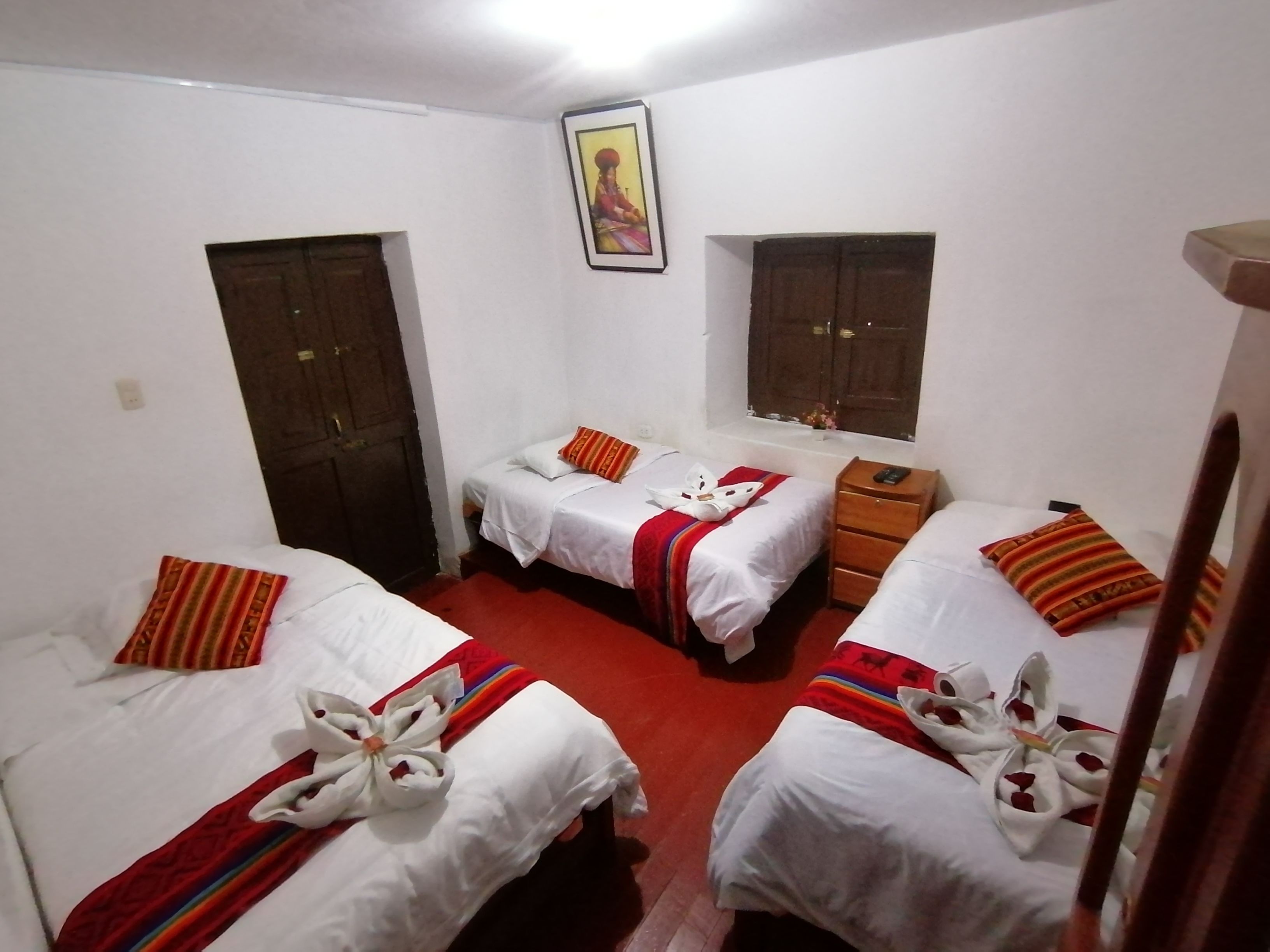 A hotel room with three single beds, each with a white duvet and red-striped pillows. There is a wooden nightstand with a phone on it, and a window with a wooden shutter.