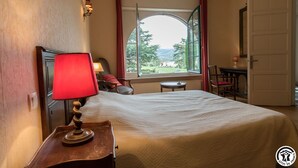 Una habitación de hotel con una cama, una lámpara roja, un ventanal con vistas a zonas verdes, una mesa con una silla y una puerta.