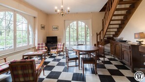 Un salón espacioso con suelo de cuadros, una mesa de comedor y un televisor. Hay ventanas grandes y una escalera.
