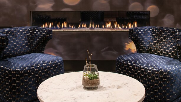 A cozy seating area with two patterned armchairs and a small table, featuring a fireplace with a visible fire. - Harlow Hotel (Portland)