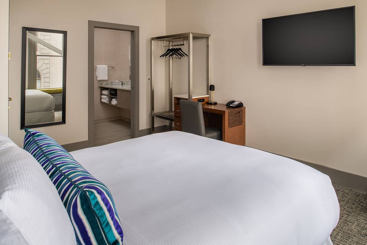Habitación de hotel con una cama grande, un escritorio, una silla, un armario, un espejo y un televisor de pantalla plana.