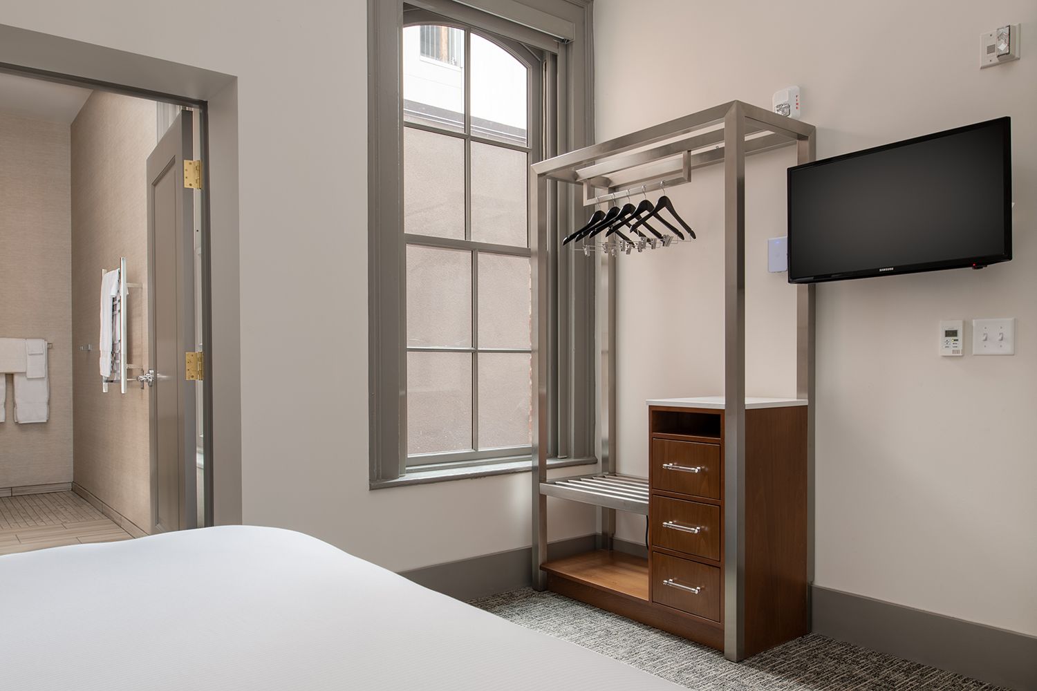A hotel room with a bed, a wardrobe with hanging clothes, a TV mounted on the wall, and a window with a view.
