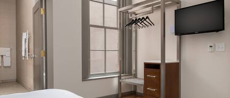 A hotel room with a bed, a wardrobe with hanging clothes, a TV mounted on the wall, and a window with a view.