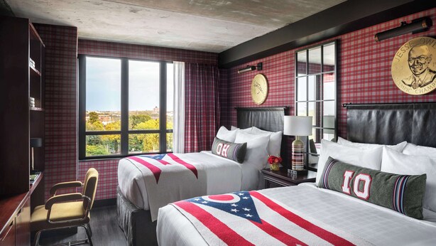 Habitación de hotel con dos camas, un escritorio, una silla y ventanas amplias con vista a la ciudad.