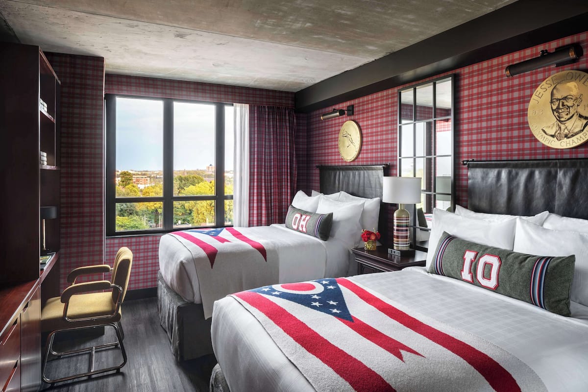 A hotel room with two beds, a desk, a chair, and large windows with a city view.