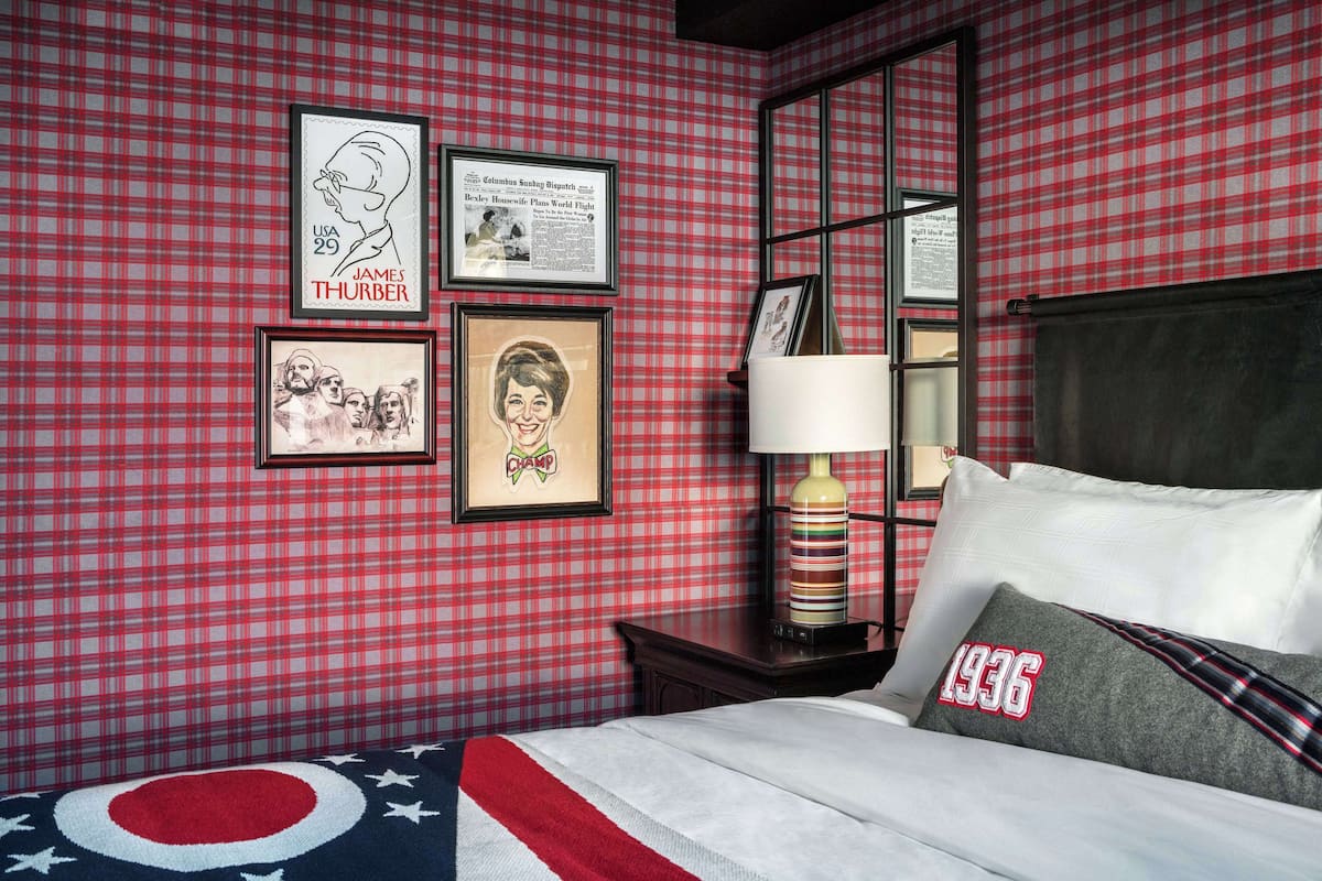 A hotel room with a checkered red and white wall, a bed with a white and red striped blanket, and a pillow with '1936' embroidered on it.