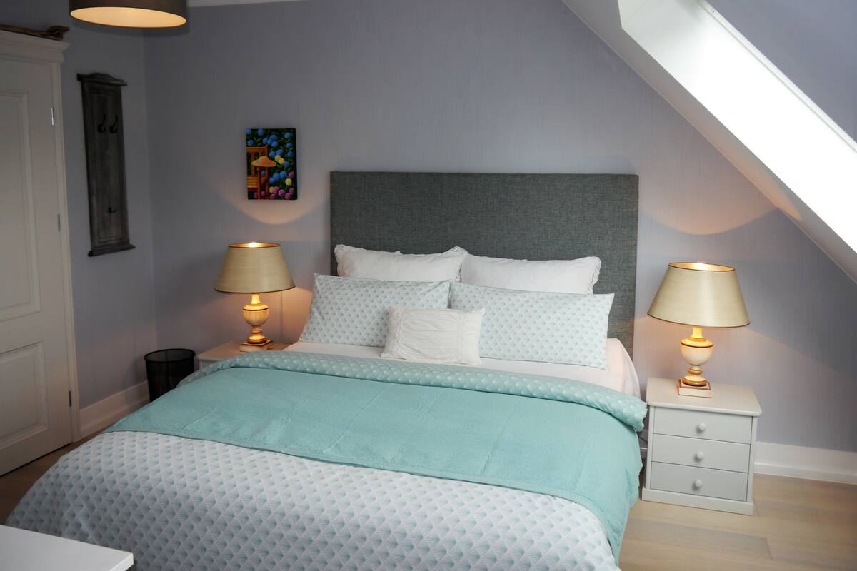 A bedroom with a bed, two bedside tables with lamps, a nightstand, a wall clock, and a painting.